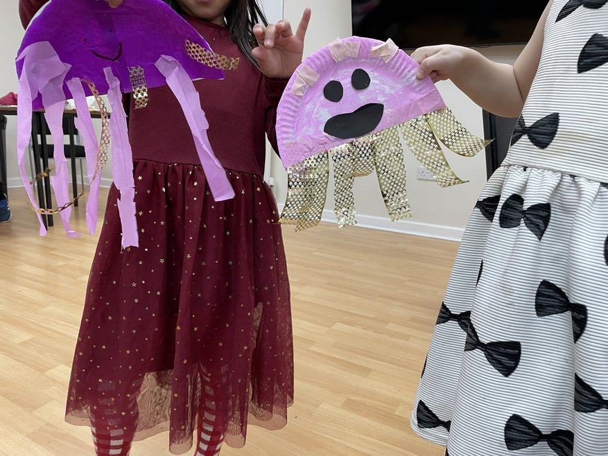 2 jellyfish created out of paper plates and strips of tissue paper. 2 girls are holding them up.