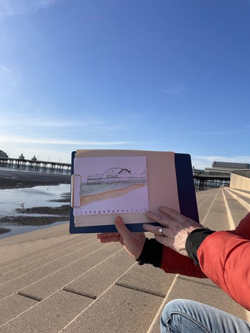 A person's hands holding up their sketch of the pier in the background to the camera