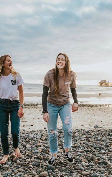 2 teenage girls laughing on the beach. It is a rocky beach with a small pier in the distance and appears to be dusk.