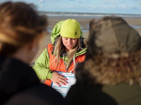 A woman wearing a lime green beanie and orange safety vest is looking down at a sheet on a clipboard. She is on a beach and is explaining the sheet to 2 people in the foreground