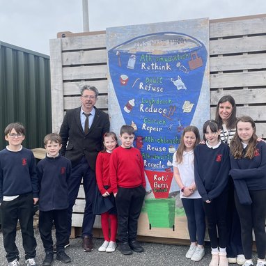 Some students and 2 adults standing in front of an outside wall with an image of a waste funnel