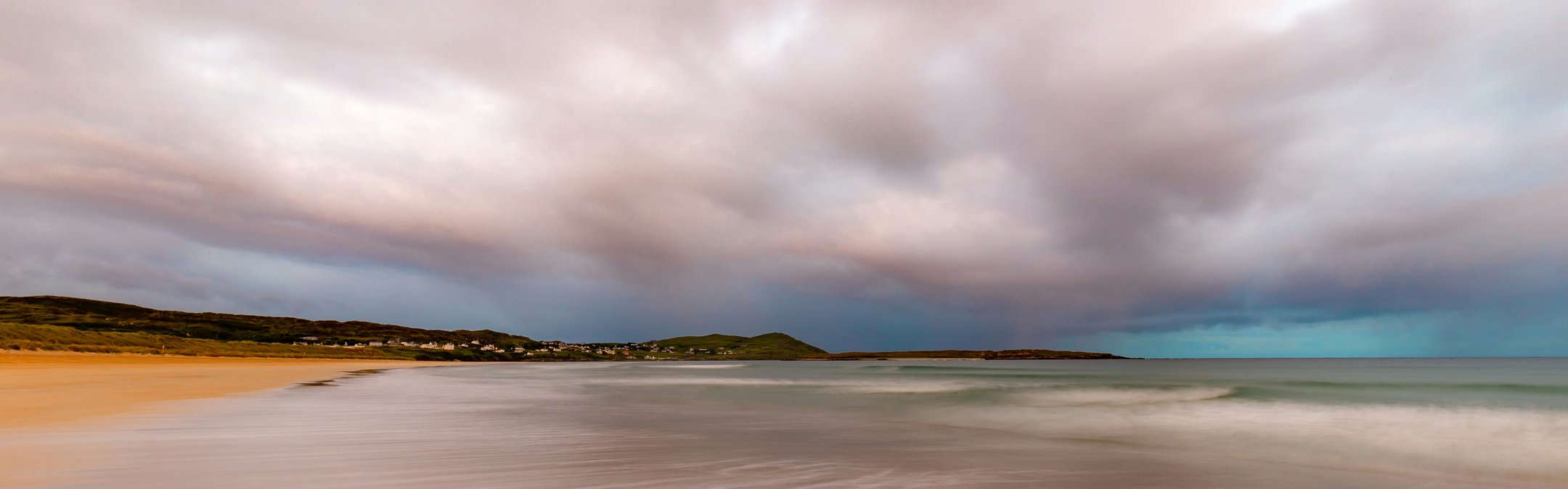 An expansive, long-exposure photograph of Narin Strand, a sandy beach, under a dramatic, overcast sky. The motion blur of the receding tide creates smooth, misty white streaks across the foreground.