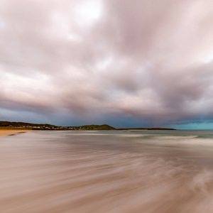 An expansive, long-exposure photograph of Narin Strand, a sandy beach, under a dramatic, overcast sky. The motion blur of the receding tide creates smooth, misty white streaks across the foreground.