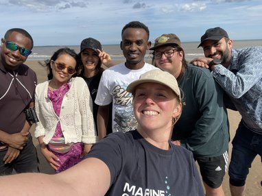 A selfie taken on a camera with 7 people posing for the camera. Behind them is a sunny beach. The girl taking hte picture is wearing a Marine Conservation Society t-shirt.