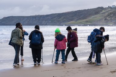 Children wearing coats and backpacks walking along the coast taking part in a beach clean. Some are holding litter pickers. Some cliffs are visible behind them.