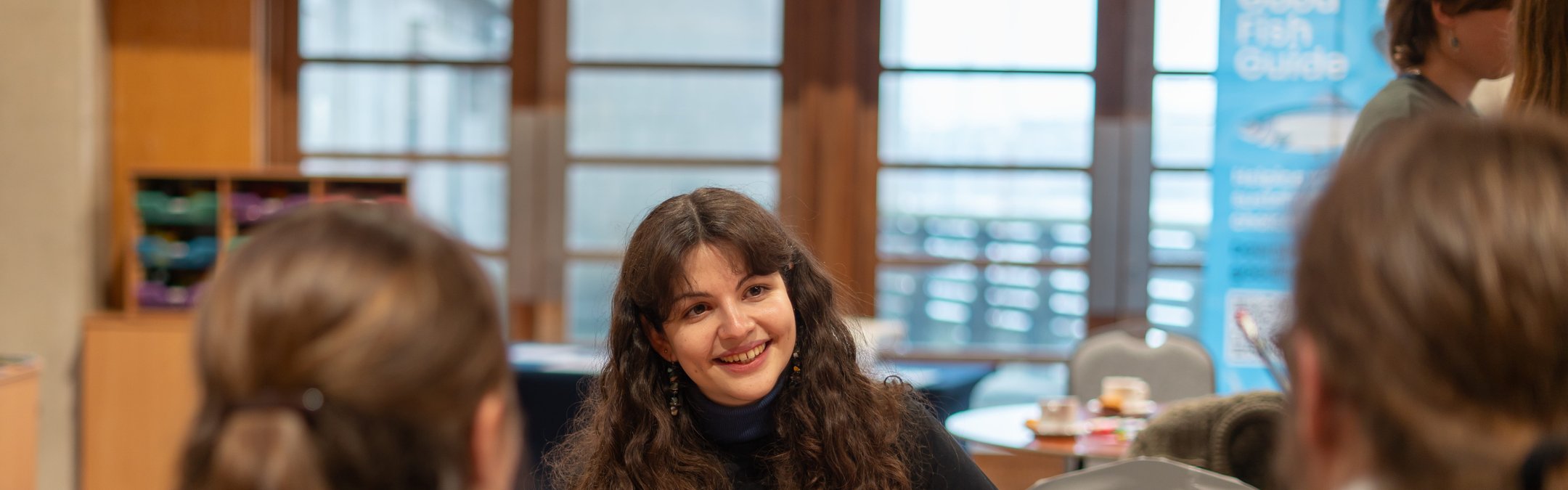 A young adult with long hair smiling while talking to 2 other people around a table. The 2 other people have their backs to the camera.