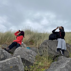 2 people sititng on a grassy hill looking upward through binoculars. There are a few big stones in front of them on the hill.