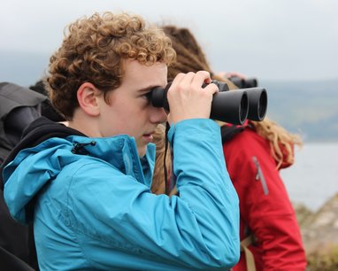 A young person looking through binoculars outside. They are wearing a light blue coat and there is someone standing behind htem wearing a red coat. In the background is the ocean.
