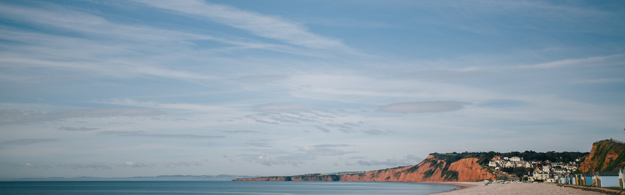 A coastal shot of Budleigh Salterton pebble beach. In the background, the distinctive red cliffs can be seen.