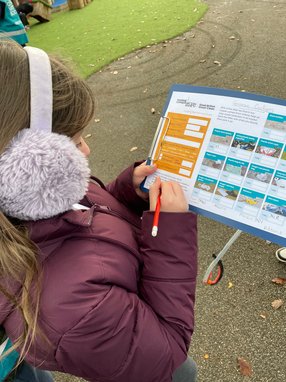 A child wearing earmuffs and a purple coat is taking part in a Source to Sea survey. She is holding a form and writing on it with a red pencil.
