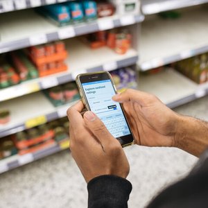 A person stands in front of the fish section in a supermarket holding a phone in their hand. On the phone screen is the Good Fish Guide species ratings page.