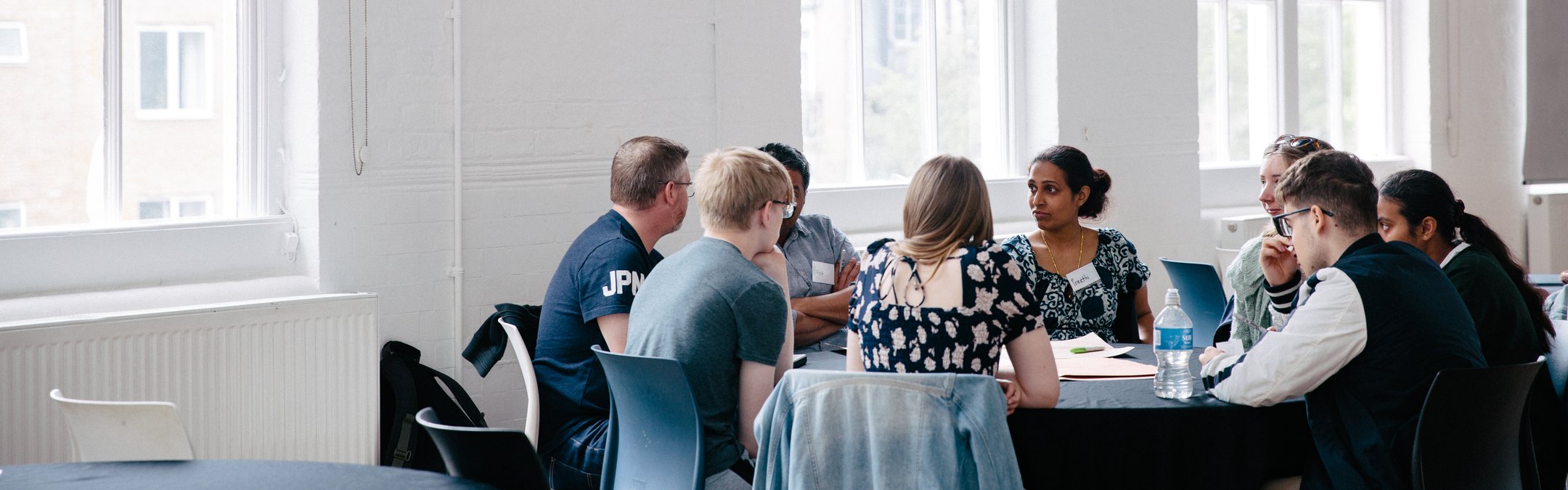A group of young adults sitting around a table. The room is white and windows are to the left and behind them