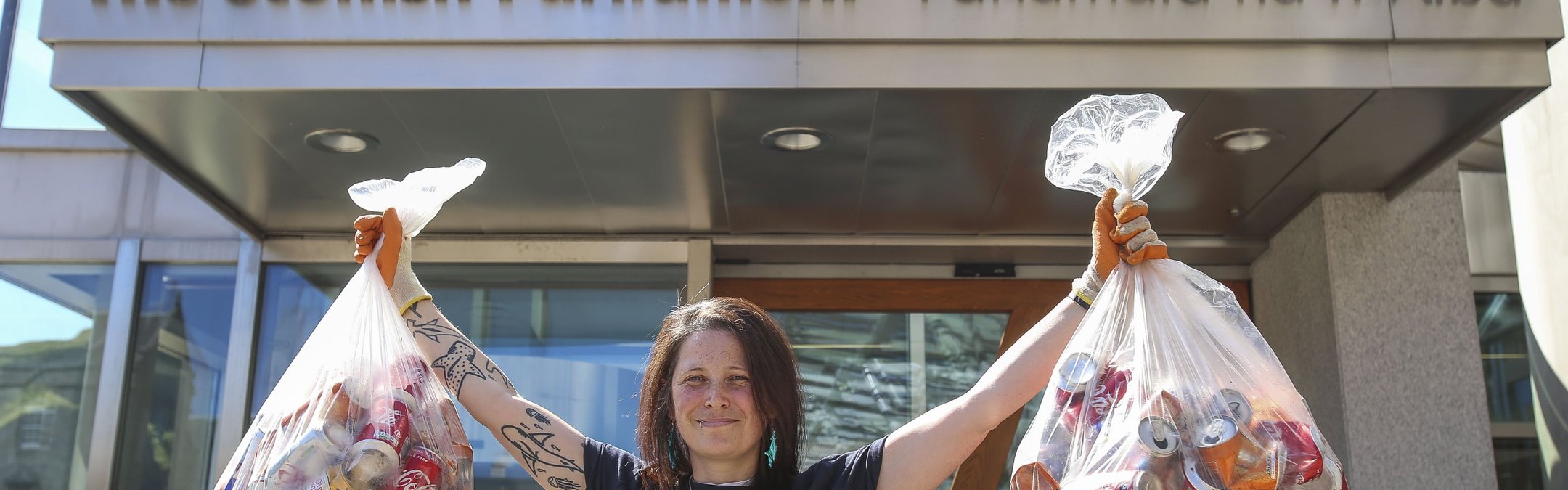 A female stands facing the camera wearing a dark blue Marine Conservation Society t-shirt. She has both arms in the air, holding two bags of plastic bottles.