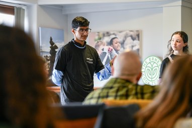 A young adult standing in front of some seated adults. He is wearing a Marine Conservation Society t-shirt and is speaking to the audience. Another young adult is standing near them listening to their speech.