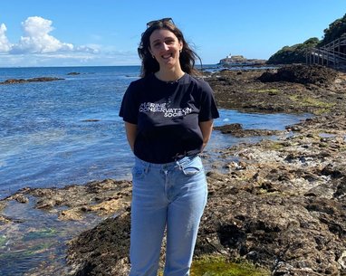 A member of Marine Conservation Society standing near some rockpools smiling at the camera. They are wearing a branding t-shirt and light blue jeans.