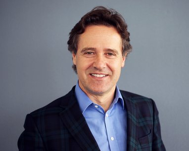 Trustee Stefano smiles against a plain grey background. It is a headshot style photo and he is wearing a blue shirt and green and blue checked jacket.