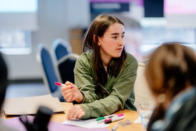 A young adult sitting at a table looking to their left away from the camera. They are holding a pen in their right hand.